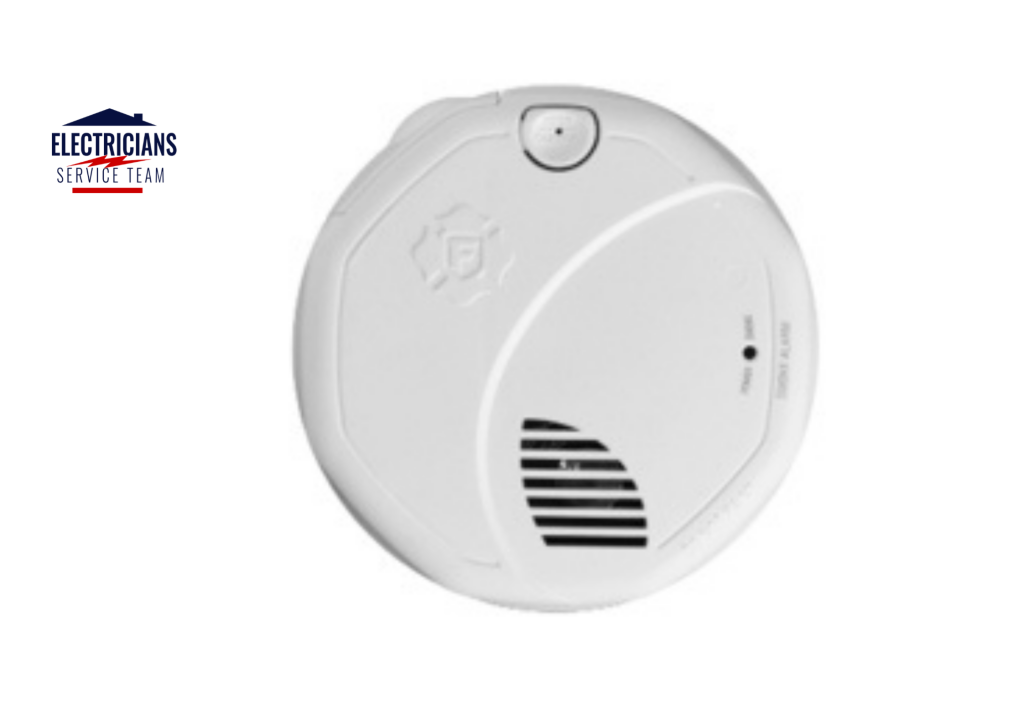 A round, white First Alert smoke detector featuring a sleek modern design. The device includes a large test/silence button at the top, a speaker grille for voice alerts at the bottom, and a small LED indicator light labeled for power and smoke status.